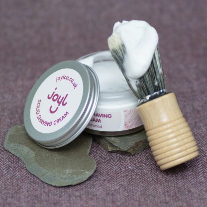 An open jar of solid shaving cream with a shaving brush lathered with cream, leaning on the jar. The background is pieces of slate on tweed fabric